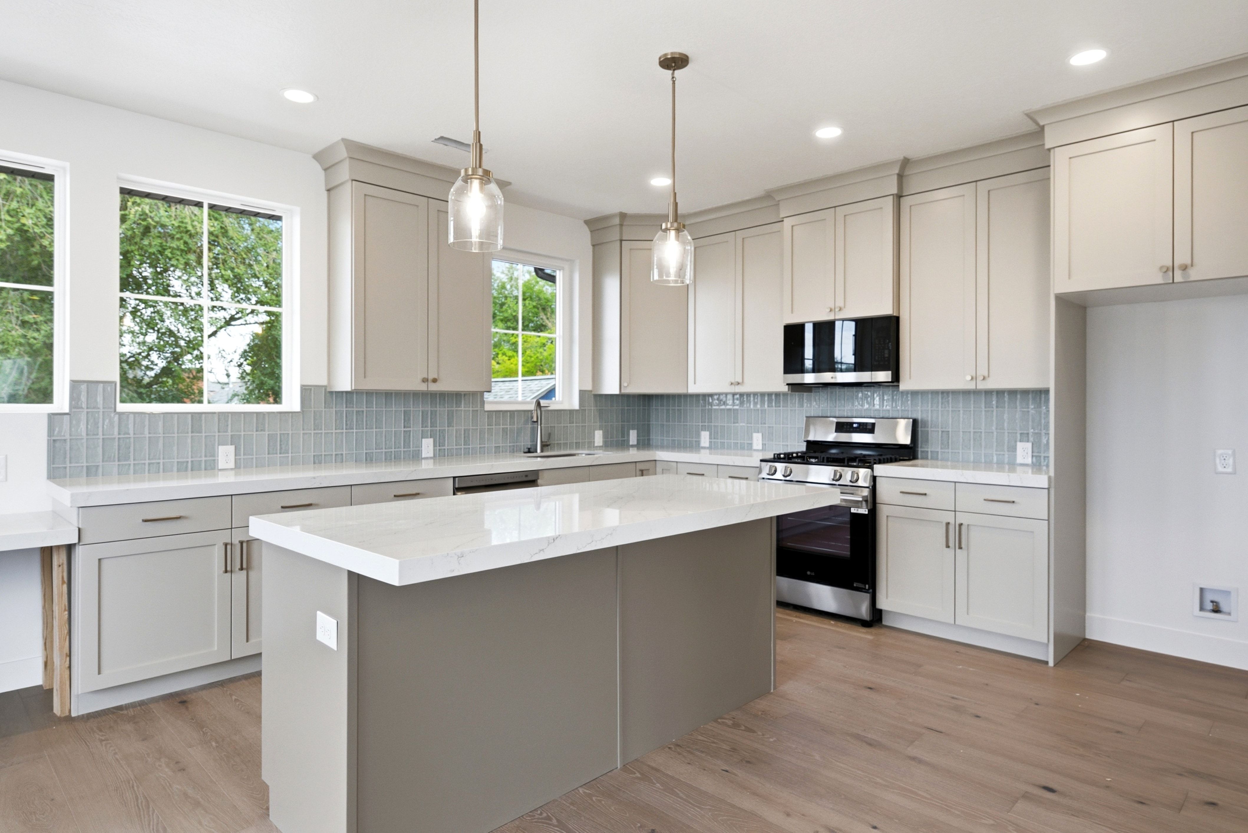 Contemporary kitchen featuring an expansive island with a white countertop, light gray cabinetry, a mosaic tile backsplash, stainless steel appliances, and wood-finish flooring