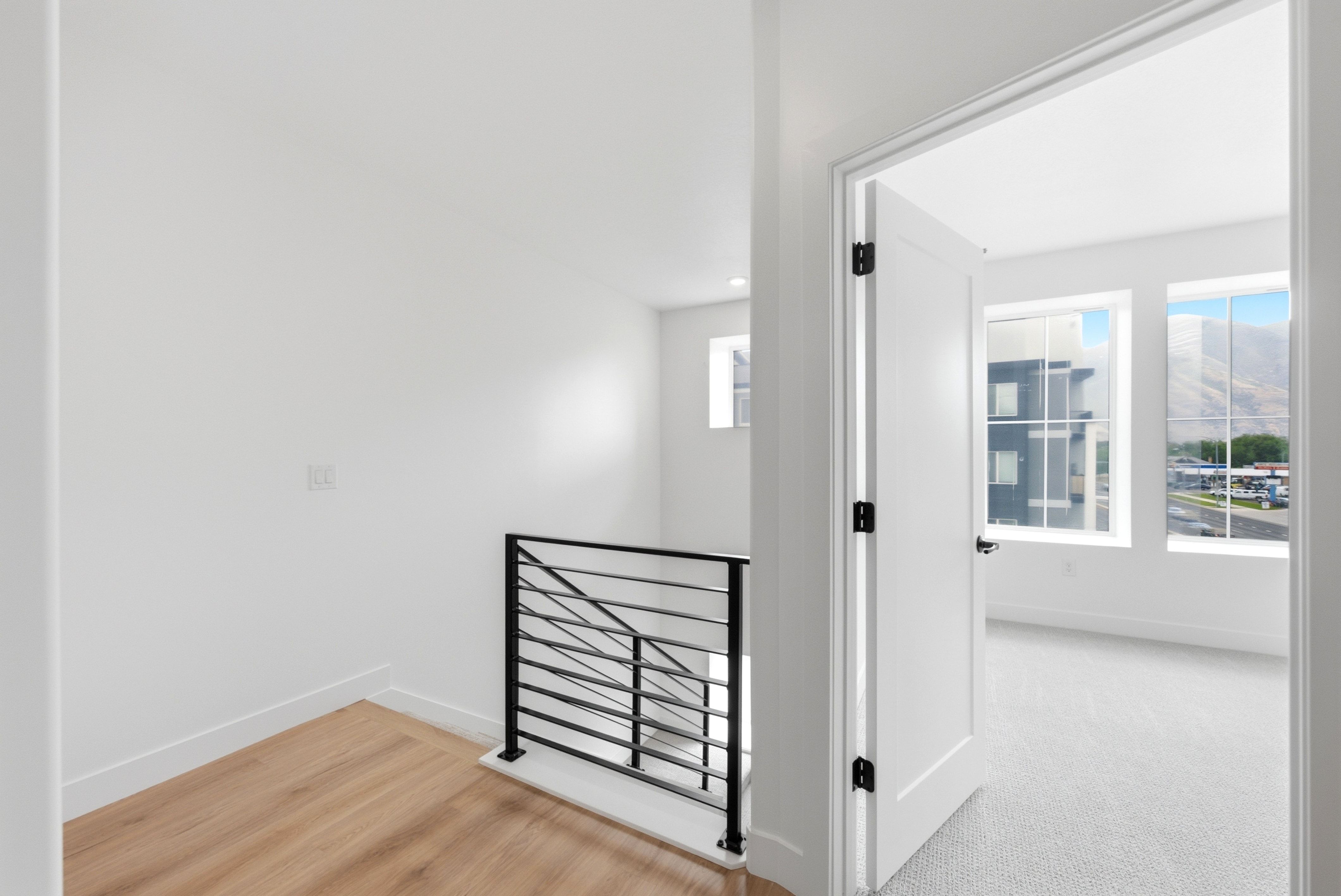 Open interior doorway leading to a room with dual windows, wood-finish flooring in the hallway, and a modern black metal stair railing