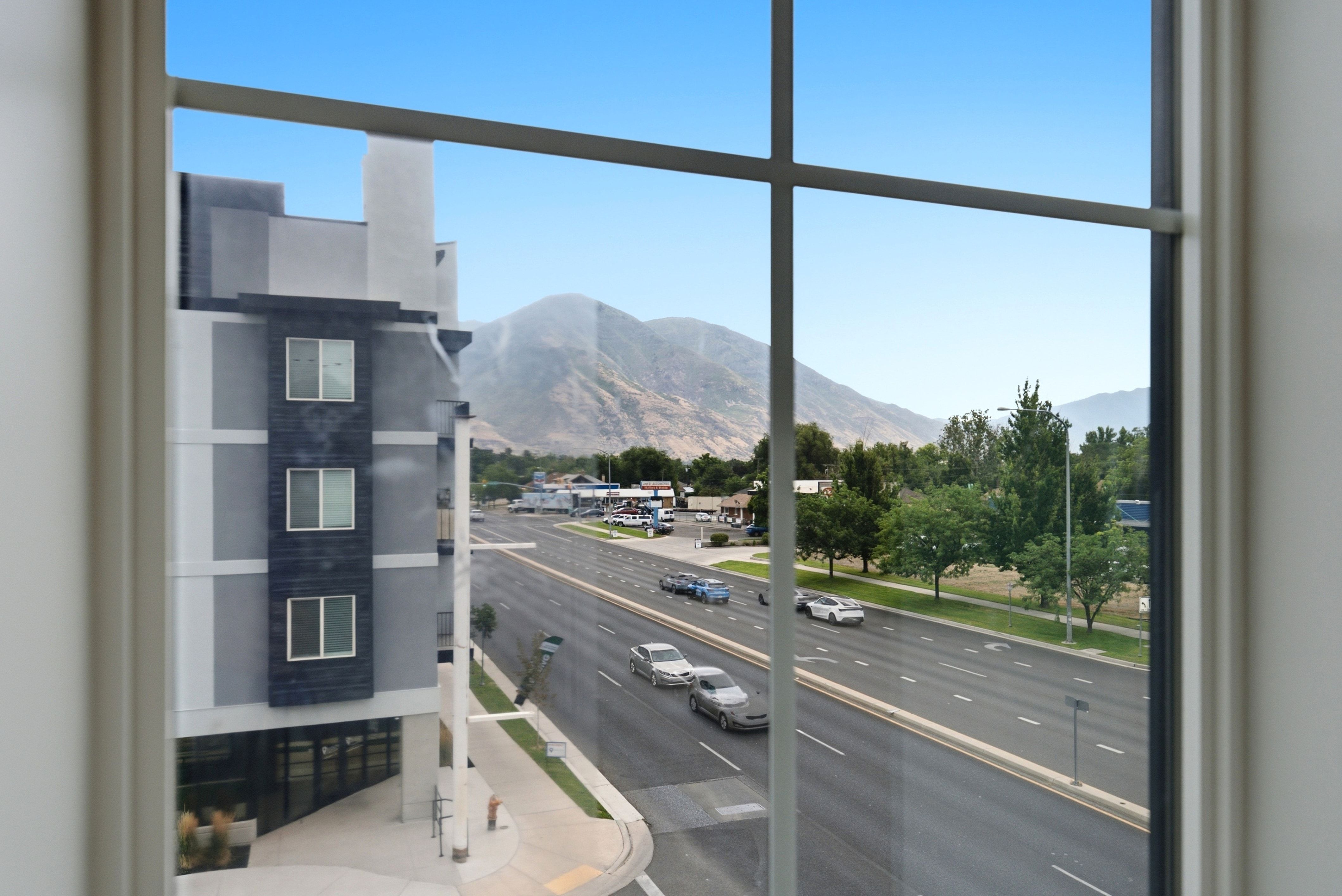 Mountain scenery framed by a large window with grid-pattern panes, showcasing a modern multi-story building, a multi-lane road, and mature trees