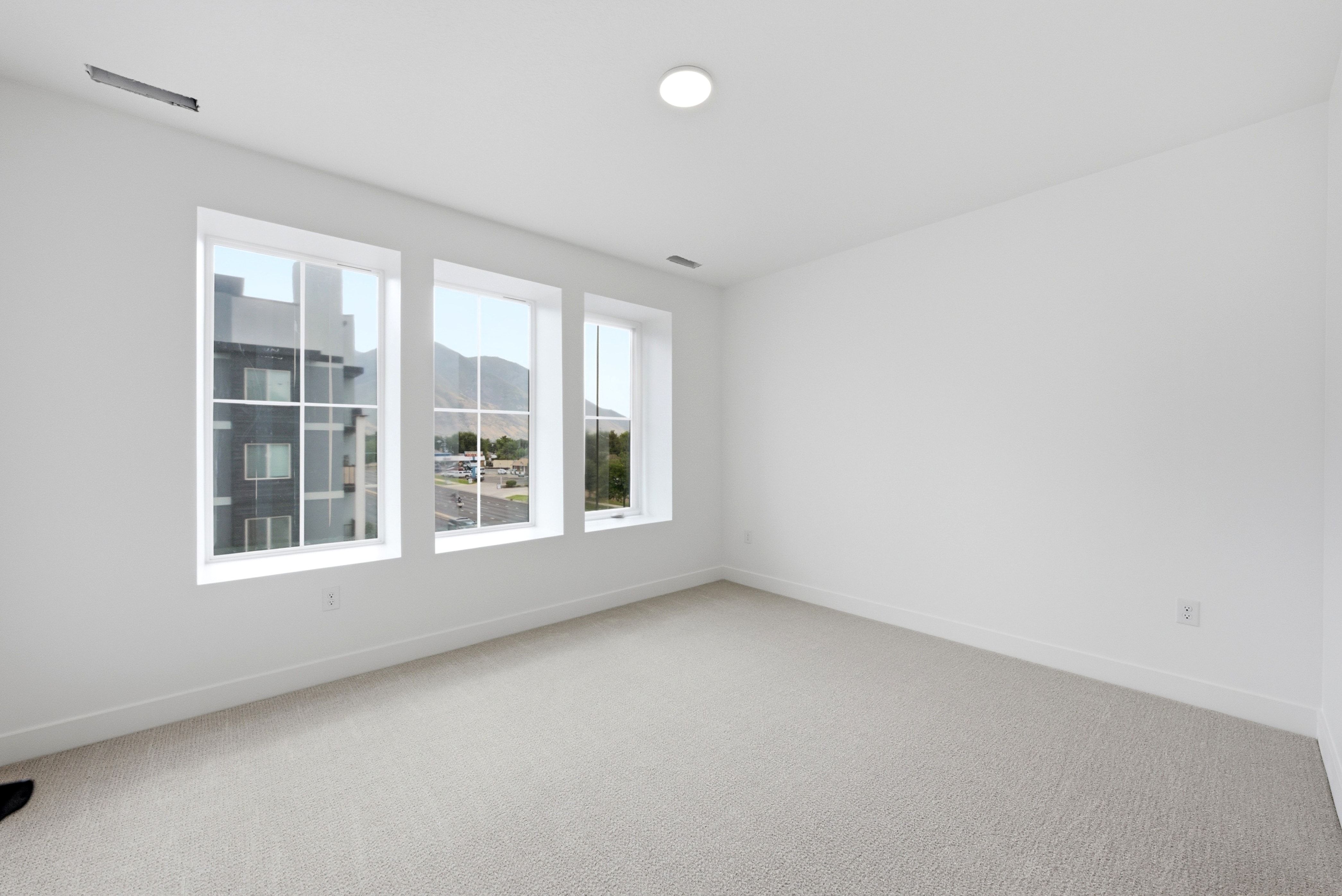 Bright interior space featuring three large windows, neutral carpeting, white walls, and a ceiling-mounted light fixture