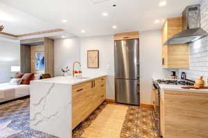 Kitchen with a peninsula, light brown cabinets, wall chimney range hood, stainless steel appliances, and light stone counters
