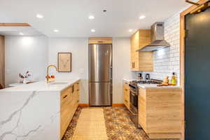 Kitchen featuring a peninsula, stainless steel appliances, modern cabinets, light stone countertops, and wall chimney exhaust hood
