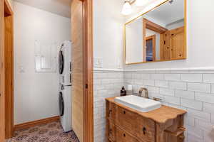 Bathroom with stacked washer / drying machine, vanity, electric panel, tile walls, and a wainscoted wall