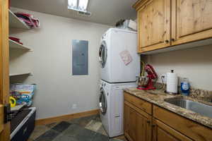 Laundry room featuring stacked washer / dryer, electric panel, cabinet space, and stone tile flooring
