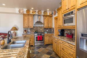 Kitchen with stainless steel appliances, wall chimney exhaust hood, brown cabinets, dark stone counters, and recessed lighting