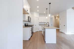 Kitchen with stainless steel appliances, light wood-style flooring, white cabinetry, a center island with sink, and decorative light fixtures