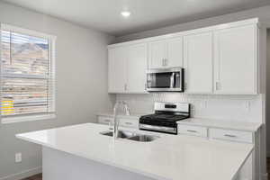Kitchen featuring appliances with stainless steel finishes, white cabinets, a center island with sink, tasteful backsplash, and light stone counters