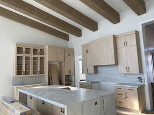 Kitchen featuring light brown cabinetry, beam ceiling, decorative backsplash, light stone countertops, and glass insert cabinets