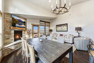Dining room with dark wood finished floors, a stone fireplace, vaulted ceiling, and a chandelier