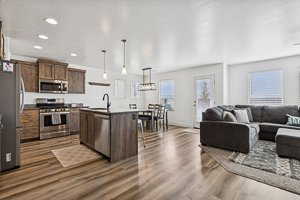 Kitchen featuring open floor plan, appliances with stainless steel finishes, an island with sink, decorative light fixtures, and dark wood-style flooring