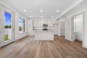 Kitchen featuring an island with sink, white cabinetry, light wood-style flooring, light stone counters, and stainless steel microwave