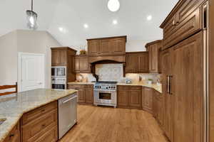 Kitchen with pendant lighting, brown cabinets, light stone counters, built in appliances, and vaulted ceiling
