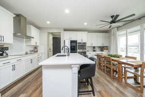 Kitchen featuring wall chimney exhaust hood, a breakfast bar, white cabinets, an island with sink, and recessed lighting