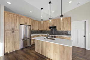 Kitchen with light brown cabinets, high end appliances, an island with sink, backsplash, and hanging light fixtures