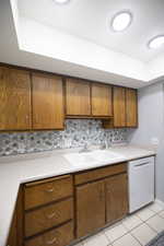 Kitchen with decorative backsplash, light countertops, brown cabinetry, and white dishwasher