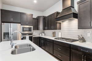 Kitchen with stainless steel appliances, custom range hood, dark brown cabinetry, tasteful backsplash, and light stone counters