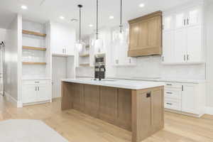 Kitchen with open shelves, decorative backsplash, white cabinets, hanging light fixtures, and a center island with sink