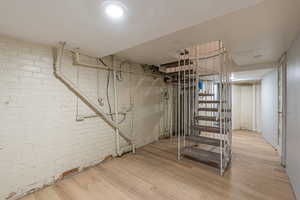 Unfinished below grade area with brick wall, light wood-type flooring, and a textured ceiling