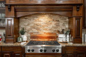 Kitchen featuring range hood, backsplash, stainless steel gas stovetop, light stone counters, and dark brown cabinets