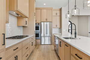 Kitchen with light brown cabinetry, appliances with stainless steel finishes, hanging light fixtures, light wood finished floors, and light stone countertops