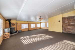 Unfurnished living room featuring a wood stove, vaulted ceiling, wood walls, ceiling fan, and carpet floors