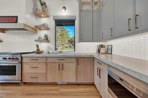 Kitchen with appliances with stainless steel finishes, custom range hood, light brown cabinetry, glass insert cabinets, and light wood-style flooring
