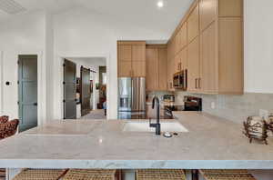 Kitchen featuring light brown cabinets, appliances with stainless steel finishes, lofted ceiling, tasteful backsplash, and light stone counters