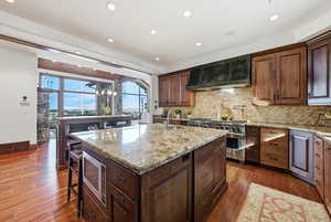 Kitchen featuring light stone countertops, dark wood finished floors, a center island with sink, backsplash, and wall chimney exhaust hood