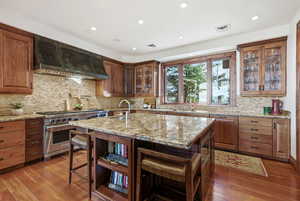 Kitchen featuring glass insert cabinets, stainless steel appliances, light stone countertops, custom range hood, and a kitchen bar
