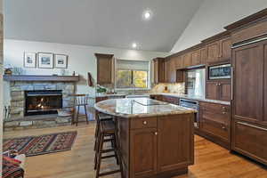 Kitchen with a fireplace, built in appliances, vaulted ceiling, light stone counters, and a breakfast bar
