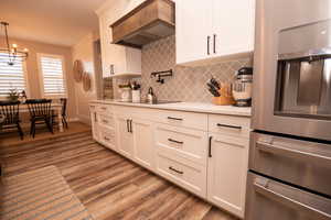 Kitchen featuring stainless steel refrigerator with ice dispenser, custom exhaust hood, light wood finished floors, a chandelier, and white cabinetry