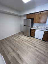Kitchen with white dishwasher, freestanding refrigerator, light wood-style floors, light stone countertops, and brown cabinets