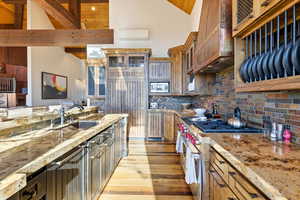 Kitchen with stainless steel appliances, custom range hood, light stone counters, light wood-style flooring, and high vaulted ceiling