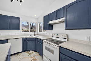 Kitchen featuring blue cabinetry, lofted ceiling, white appliances, light marble finish floors, and light countertops