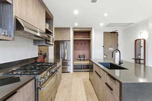 Kitchen with appliances with stainless steel finishes, modern cabinets, under cabinet range hood, light brown cabinets, and dark stone counters