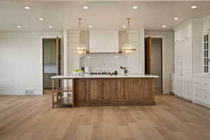 Kitchen with a large island with sink, decorative light fixtures, light wood-style floors, brown cabinetry, and white cabinets