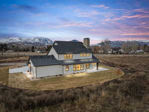 Back of house at dusk with a patio, a mountain view, a chimney, a lawn, and roof with shingles
