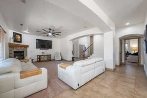 Living area with arched walkways, ceiling fan, a stone fireplace, stairs, and light tile patterned floors