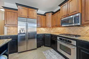Kitchen with appliances with stainless steel finishes, backsplash, light stone counters, light tile patterned flooring, and dark cabinetry