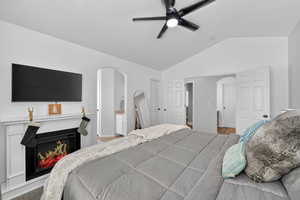 Bedroom with vaulted ceiling, a lit fireplace, a ceiling fan, and ensuite bathroom