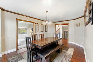 Dining area with dark wood-style floors, a wainscoted wall, ornamental molding, a chandelier, and stairs