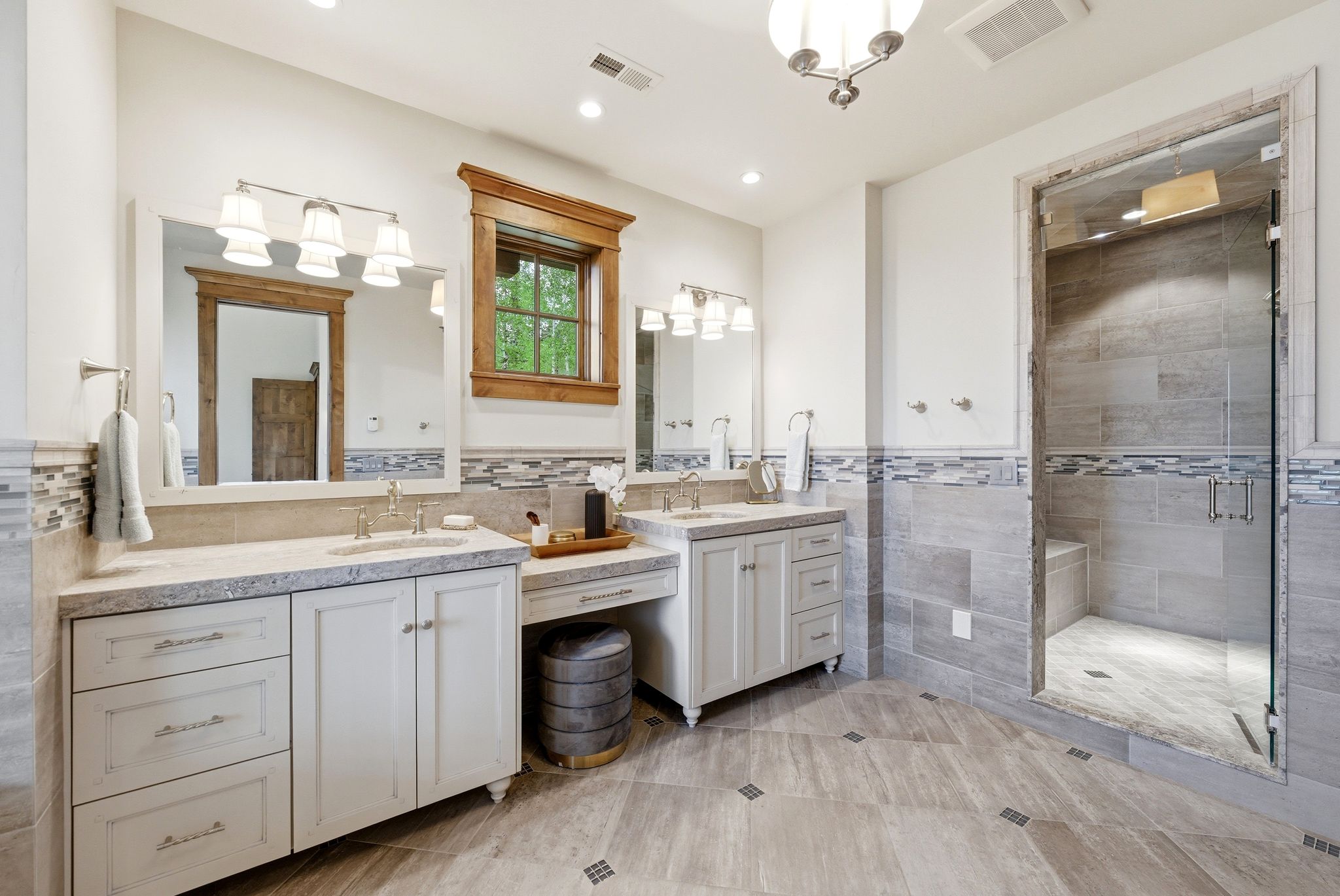 Spacious bathroom featuring a large glass-enclosed shower with a built-in bench, dual vanities with light-toned countertops, and a window framed in natural wood