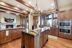 Kitchen featuring stainless steel double oven, a kitchen island, a stone fireplace, beamed ceiling, and brown cabinetry