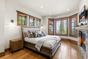 Spacious room featuring wood-finish flooring, a stone fireplace with hearth, recessed lighting, and multiple windows with wood trim and plantation shutters