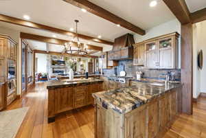 Rustic-inspired kitchen featuring wood-finish flooring, exposed ceiling beams, custom wood cabinetry, stone countertops, and a built-in range with a copper-tone hood