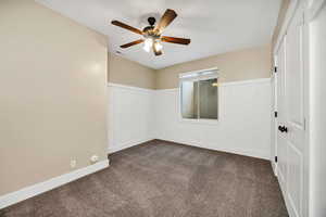 Unfurnished bedroom featuring a decorative wall, a wainscoted wall, dark carpet, and a ceiling fan
