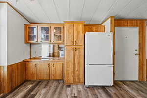 Kitchen featuring freestanding refrigerator, glass insert cabinets, wooden walls, dark wood-style floors, and brown cabinetry