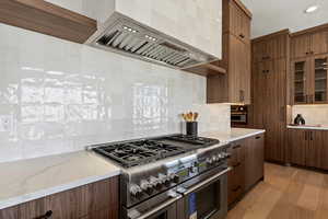 Kitchen featuring custom range hood, double oven range, light stone counters, decorative backsplash, and modern cabinets
