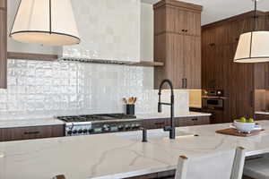 Kitchen with hanging light fixtures, light stone counters, wall chimney range hood, decorative backsplash, and oven