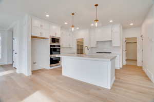 Kitchen featuring white cabinets, decorative backsplash, light wood finished floors, decorative light fixtures, and a kitchen island with sink
